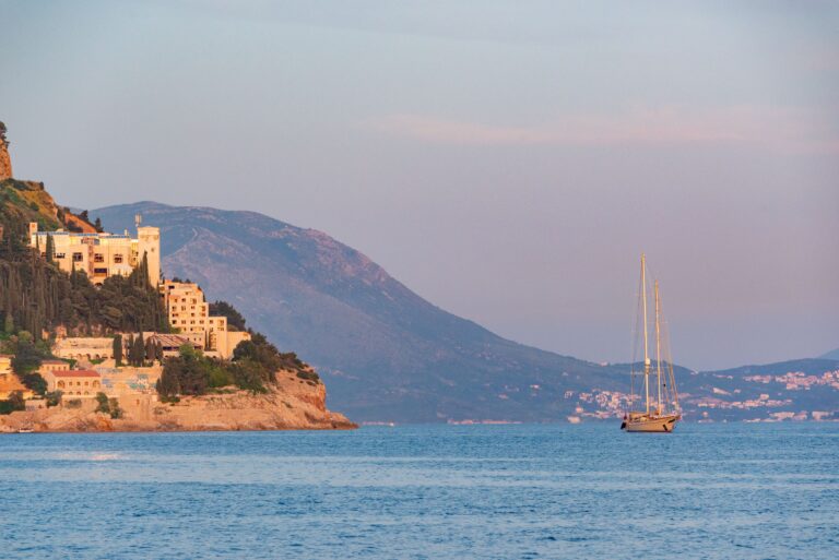 A stunning sunset view of the Dubrovnik coast with a sailboat in the Adriatic Sea, highlighting the serene beauty.