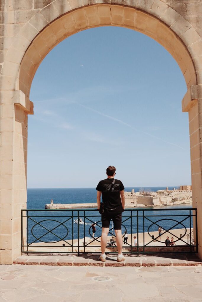 A man stands under an arch, overlooking the sea in Valletta, Malta on a sunny day.